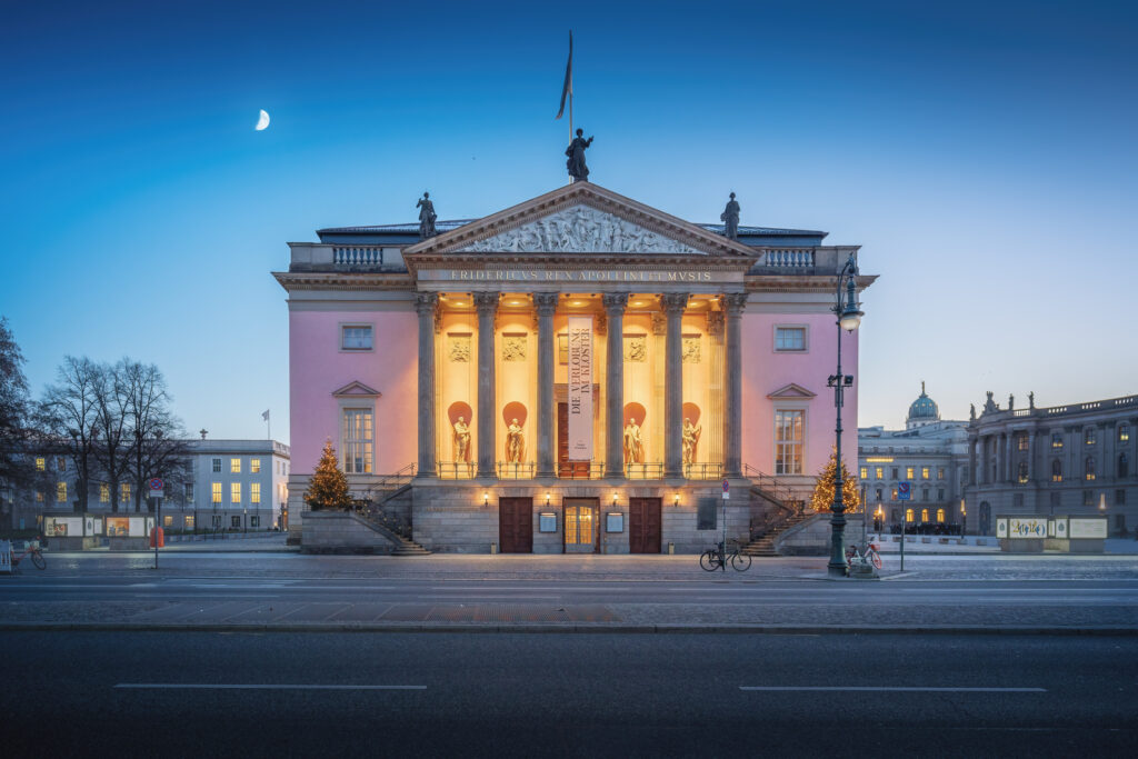 Berliner Staatsoper bei Nacht, klassizistische Fassade warm beleuchtet, menschenleerer Platz.