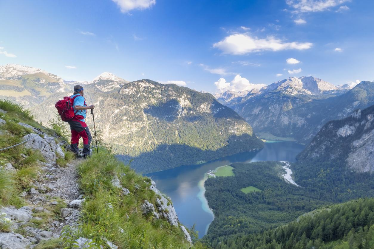 Wanderer steht auf einem schmalen Bergpfad und blickt auf den tiefblauen Königssee im Nationalpark Berchtesgaden, umgeben von hohen Alpenbergen.