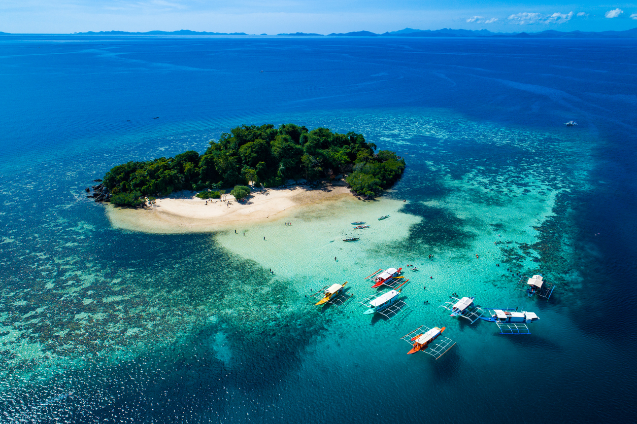 Luftaufnahme einer kleinen, von Korallenriffen umgebenen Insel mit weißem Sandstrand und mehreren traditionellen Auslegerbooten vor der Küste der Philippinen.