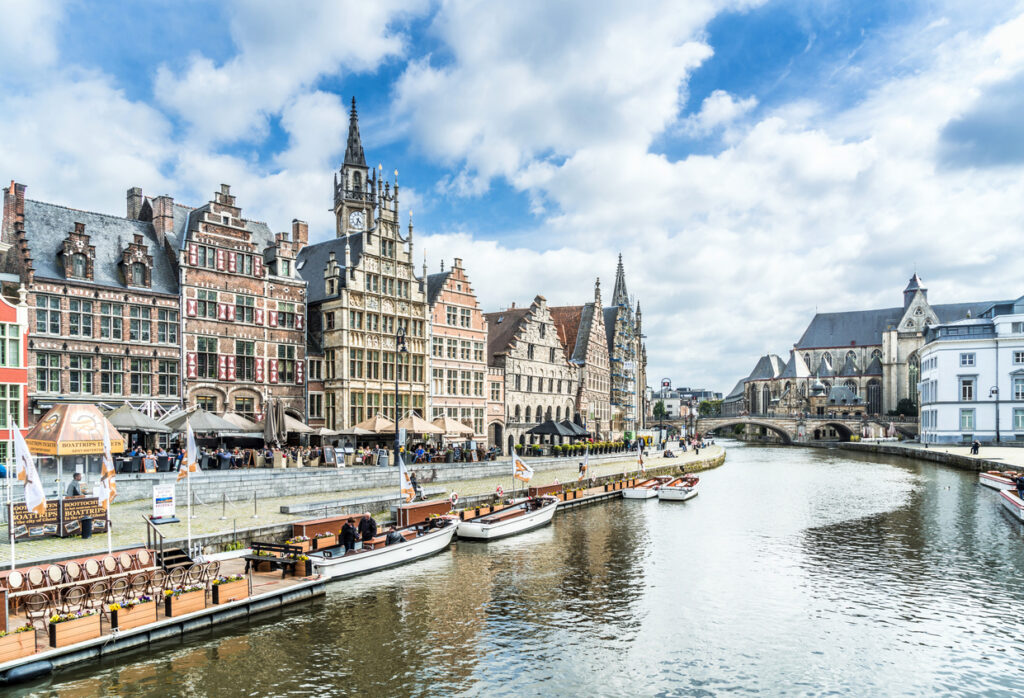Panoramablick auf die historischen Giebelhäuser am Graslei und Korenlei in Gent, Belgien, mit Booten am Kai und der St.-Nikolaus-Kirche im Hintergrund.