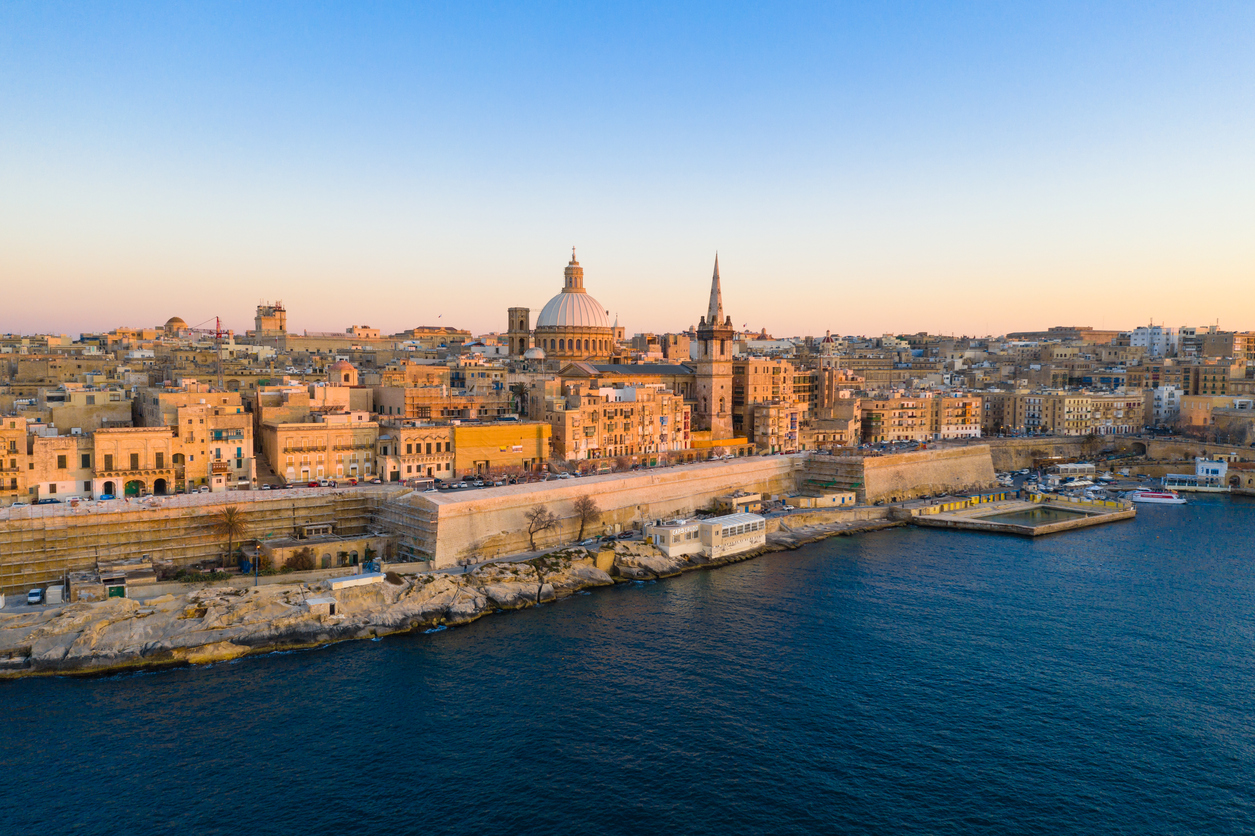 Blick auf die Altstadt von Valletta, Malta, mit der Kuppel der Karmeliterkirche und dem Turm der St. Paul’s Pro-Cathedral bei goldenem Sonnenuntergang, aufgenommen vom Meer aus.