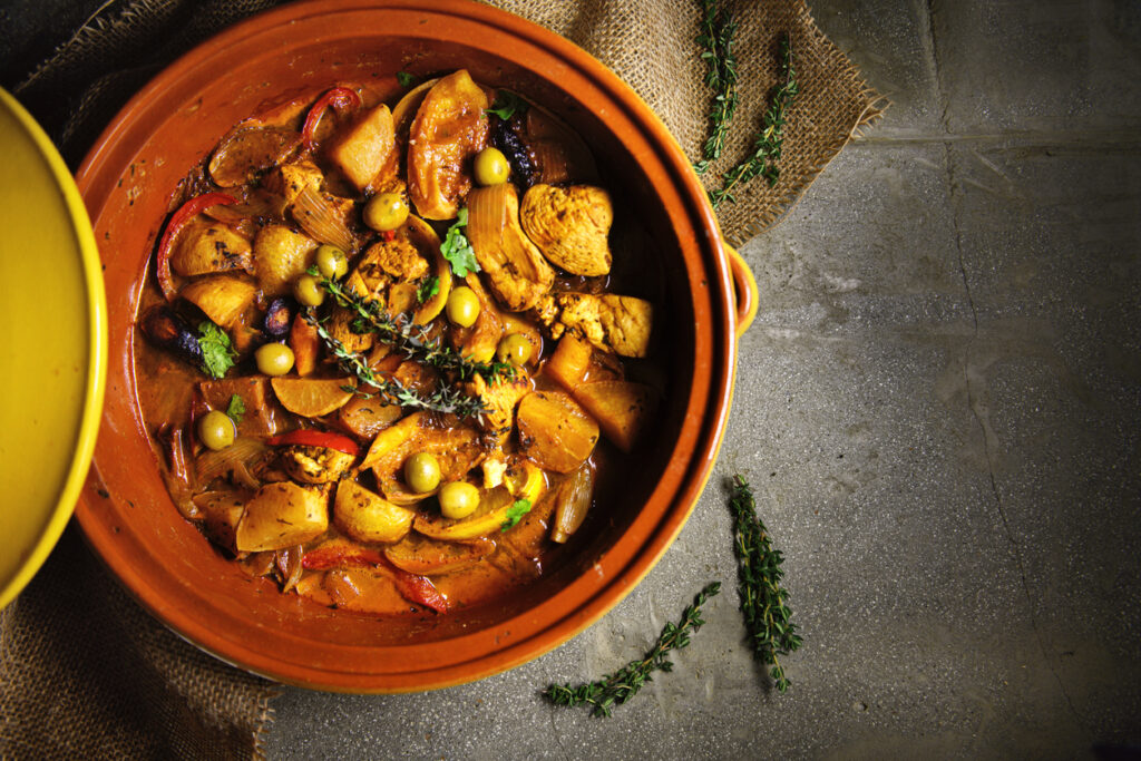 Obenansicht einer marokkanischen Tajine mit Hühnerfleisch, Kartoffeln, Karotten, Oliven, Zwiebeln und frischen Kräutern in einer traditionellen Tonform, auf einem rustikalen Untergrund serviert.