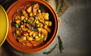 Obenansicht einer marokkanischen Tajine mit Hühnerfleisch, Kartoffeln, Karotten, Oliven, Zwiebeln und frischen Kräutern in einer traditionellen Tonform, auf einem rustikalen Untergrund serviert.