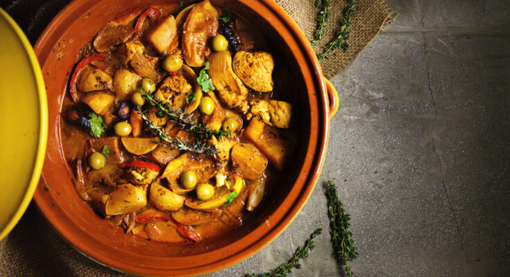 Obenansicht einer marokkanischen Tajine mit Hühnerfleisch, Kartoffeln, Karotten, Oliven, Zwiebeln und frischen Kräutern in einer traditionellen Tonform, auf einem rustikalen Untergrund serviert.