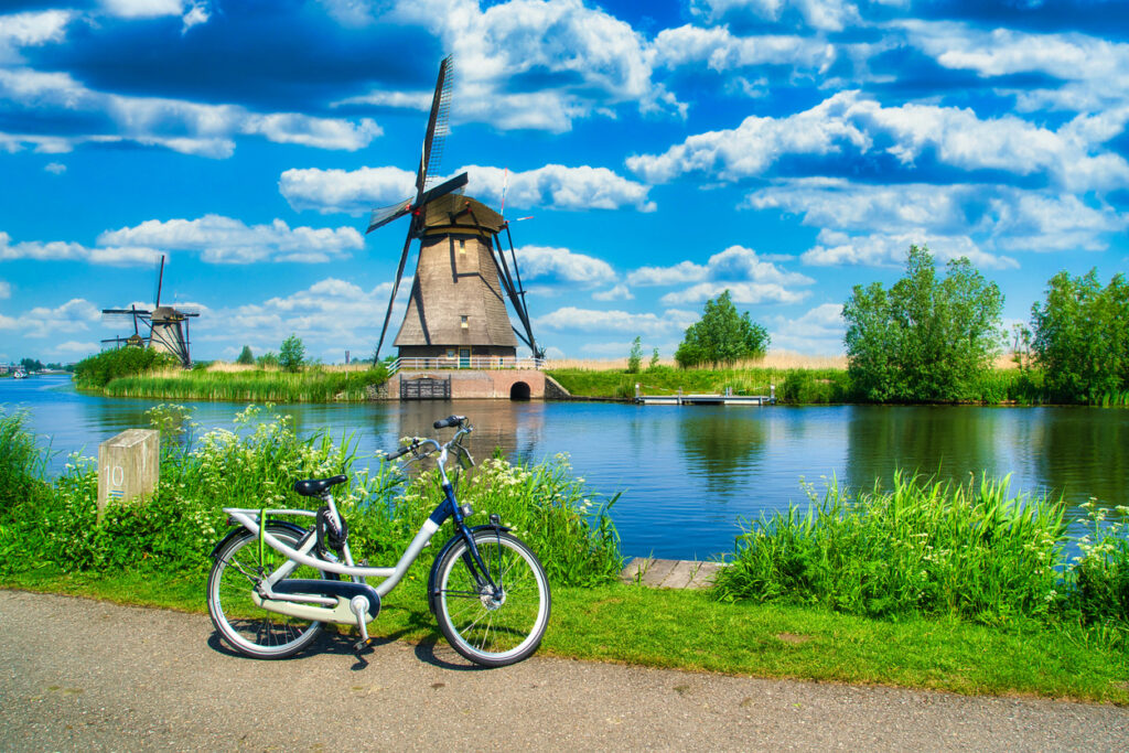 Fahrrad am Ufer eines Kanals mit Blick auf die historischen Windmühlen von Kinderdijk unter blauem Himmel mit Wolken.