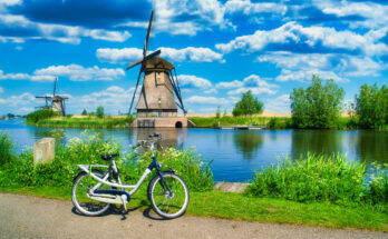 Fahrrad am Ufer eines Kanals mit Blick auf die historischen Windmühlen von Kinderdijk unter blauem Himmel mit Wolken.