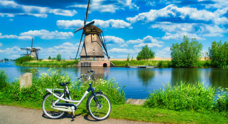 Fahrrad am Ufer eines Kanals mit Blick auf die historischen Windmühlen von Kinderdijk unter blauem Himmel mit Wolken.
