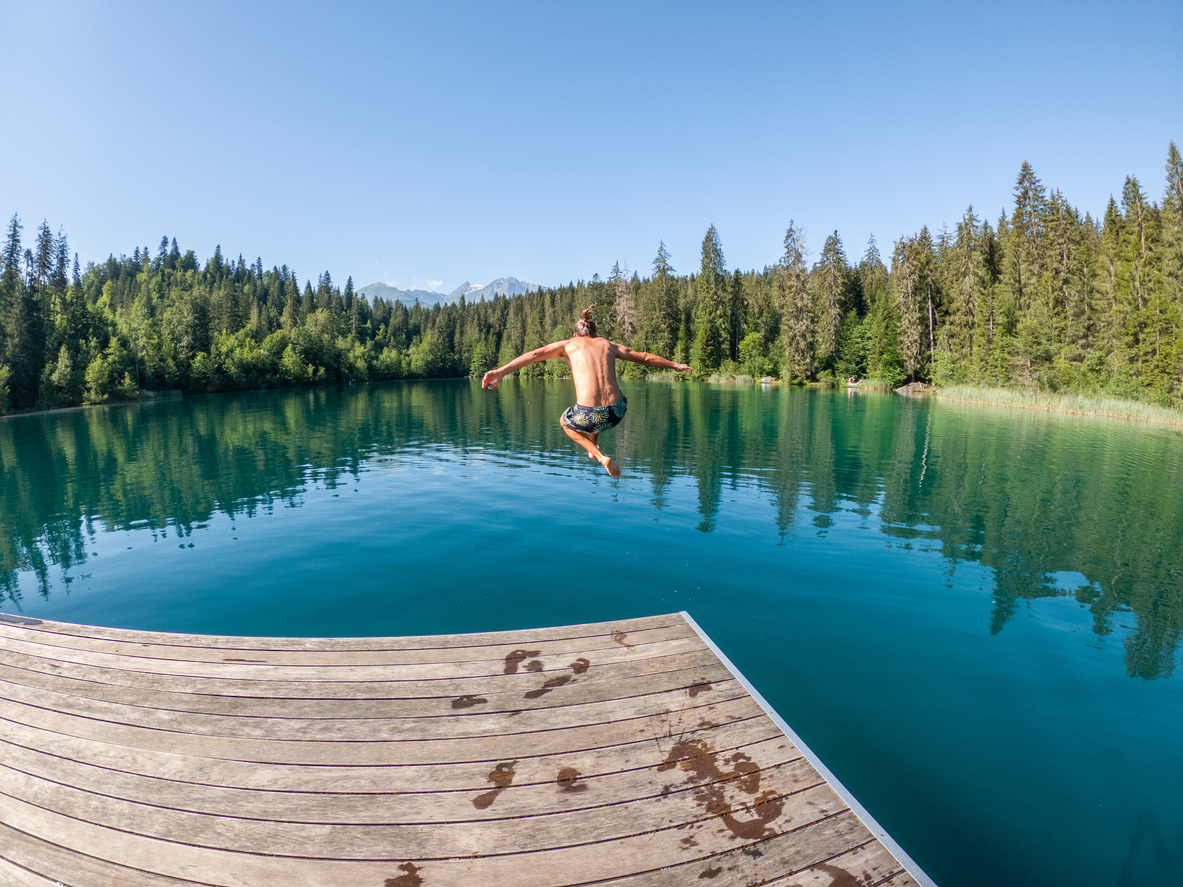 Person springt von einem Holzsteg in das klare, türkisfarbene Wasser eines von Wäldern umgebenen Schweizer Sees.