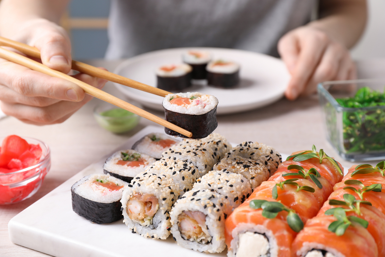 Nahaufnahme verschiedener Sushi-Sorten mit Maki, Inside-Out-Rolls mit Sesam und Lachs-Rolls, serviert auf einem weißen Teller, eine Person hält ein Stück mit Essstäbchen.