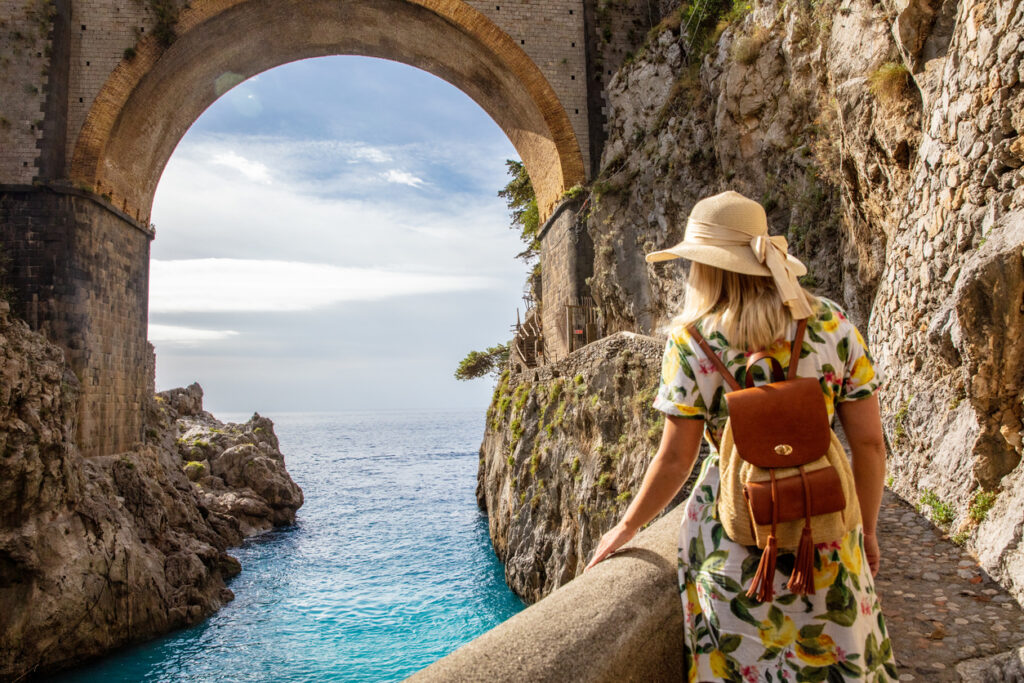 Reisende mit Sommerkleid und Strohhut blickt auf das türkisfarbene Meer unter der Steinbrücke von Furore an der Amalfiküste in Italien.