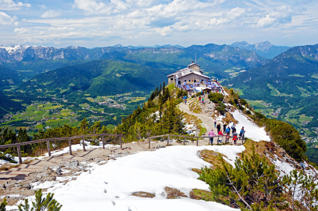 Wanderer auf dem verschneiten Pfad zum Kehlsteinhaus am Obersalzberg in Berchtesgaden, umgeben von den Alpen und einem weiten Blick ins grüne Tal.