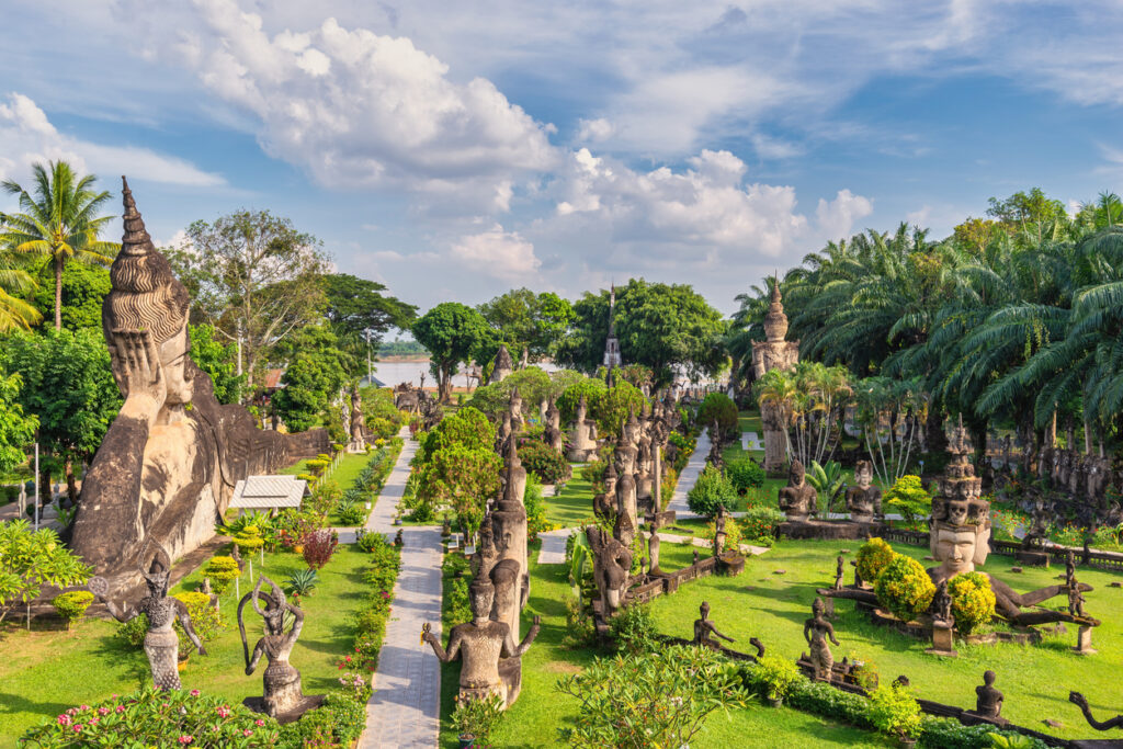 Panoramablick auf den Buddha-Park in Laos mit zahlreichen buddhistischen und hinduistischen Steinskulpturen, umgeben von tropischer Vegetation und gepflegten Wegen unter blauem Himmel.