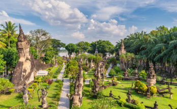 Panoramablick auf den Buddha-Park in Laos mit zahlreichen buddhistischen und hinduistischen Steinskulpturen, umgeben von tropischer Vegetation und gepflegten Wegen unter blauem Himmel.