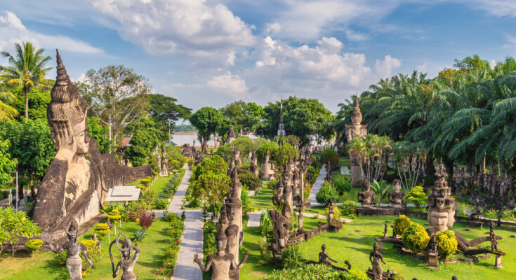 Panoramablick auf den Buddha-Park in Laos mit zahlreichen buddhistischen und hinduistischen Steinskulpturen, umgeben von tropischer Vegetation und gepflegten Wegen unter blauem Himmel.