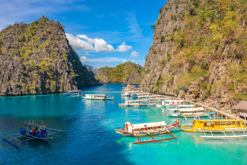Mehrere Auslegerboote liegen im klaren, türkisfarbenen Wasser zwischen steilen Kalksteinfelsen in Coron, Philippinen.
