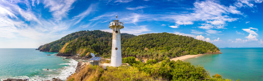 Panoramablick auf den weißen Leuchtturm im Mu Ko Lanta Nationalpark auf Koh Lanta, Thailand, mit grün bewaldeten Hügeln, Sandstrand und türkisblauem Meer.