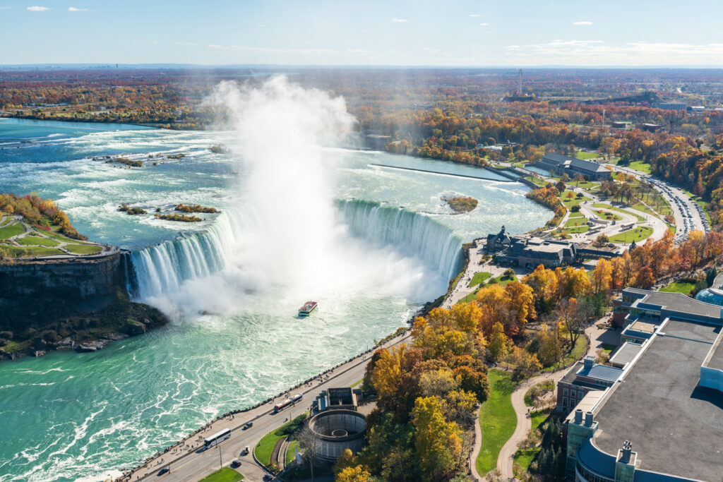 Luftaufnahme der Niagarafälle mit den Horseshoe Falls, bei denen gewaltige Wassermassen in die Tiefe stürzen, umgeben von herbstlich gefärbten Bäumen und einer Bootstour im Nebel.
