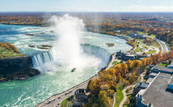 Luftaufnahme der Niagarafälle mit den Horseshoe Falls, bei denen gewaltige Wassermassen in die Tiefe stürzen, umgeben von herbstlich gefärbten Bäumen und einer Bootstour im Nebel.