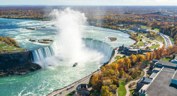 Luftaufnahme der Niagarafälle mit den Horseshoe Falls, bei denen gewaltige Wassermassen in die Tiefe stürzen, umgeben von herbstlich gefärbten Bäumen und einer Bootstour im Nebel.