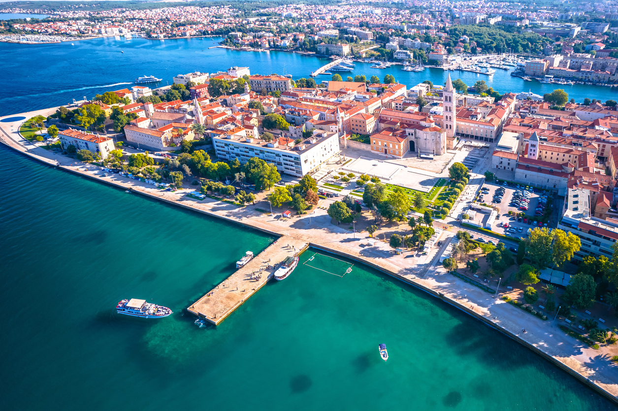 Vogelperspektive auf die Altstadt von Zadar in Kroatien, umgeben von türkisblauem Wasser, mit historischen Gebäuden, Kirchturm und Hafenanlagen.