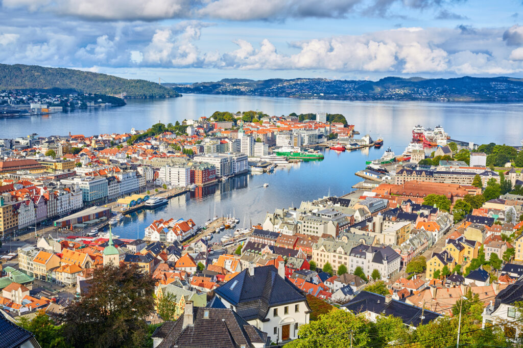 Panorama der norwegischen Stadt Bergen mit Blick auf den Hafen, farbenfrohe Häuser von Bryggen, moderne Gebäude und umgebende Fjorde unter blauem Himmel.