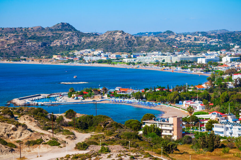Panoramablick auf die Küstenlinie von Rhodos in Griechenland mit langen Sandstränden, türkisblauem Meer, Hotels und Resorts, grünen Bäumen sowie umliegenden Bergen im Hintergrund.