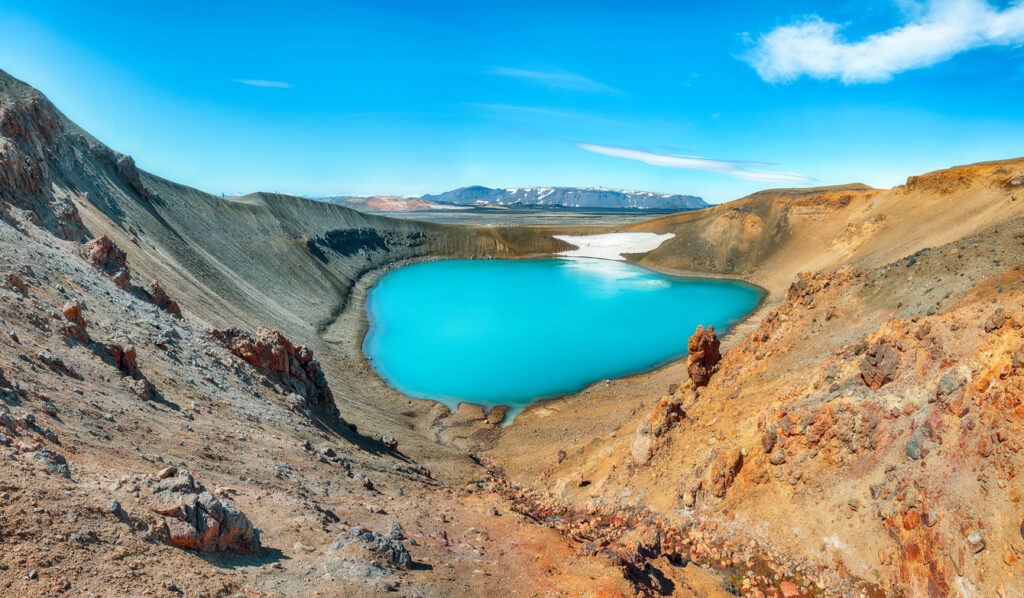 Panoramaaufnahme des türkisblauen Víti-Kratersees im Krafla-Vulkansystem, umgeben von rötlich-braunen Lavafeldern und schroffen Berghängen in Island.