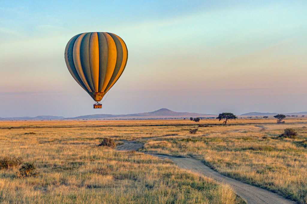 Heißluftballon schwebt über der weiten Savannenlandschaft des Serengeti-Nationalparks bei Sonnenaufgang.