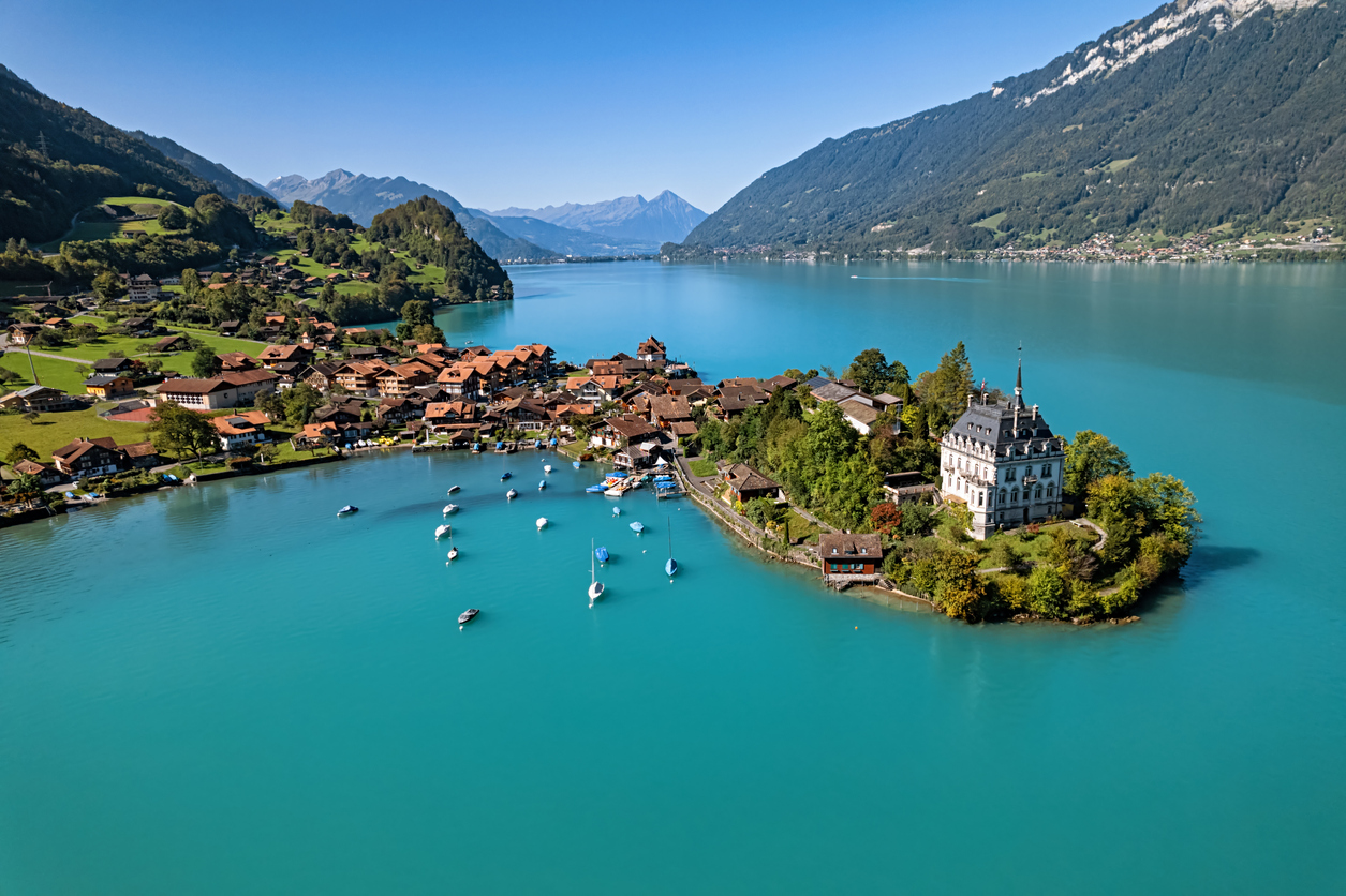 Luftaufnahme eines Schweizer Dorfes mit Segelbooten, Schloss und türkisblauem Brienzersee, umgeben von Bergen.