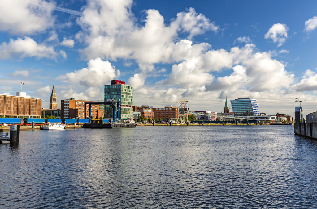 Blick über die Kieler Förde mit modernen Gebäuden, Hafenanlagen und dem Rathausturm im Hintergrund bei blauem Himmel.