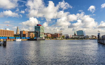Blick über die Kieler Förde mit modernen Gebäuden, Hafenanlagen und dem Rathausturm im Hintergrund bei blauem Himmel.