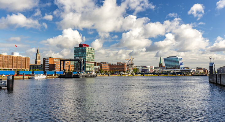 Blick über die Kieler Förde mit modernen Gebäuden, Hafenanlagen und dem Rathausturm im Hintergrund bei blauem Himmel.