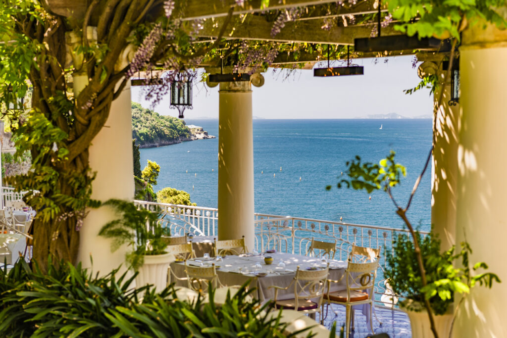 Romantische Restaurantterrasse mit Blick auf das tiefblaue Meer der Amalfiküste, umgeben von Pflanzen und überdacht von einer blühenden Pergola.