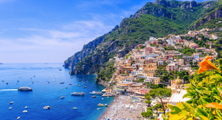 Luftaufnahme von Positano an der Amalfiküste in Italien mit bunten Häusern, Strand, Sonnenschirmen und Booten im türkisblauen Mittelmeer.