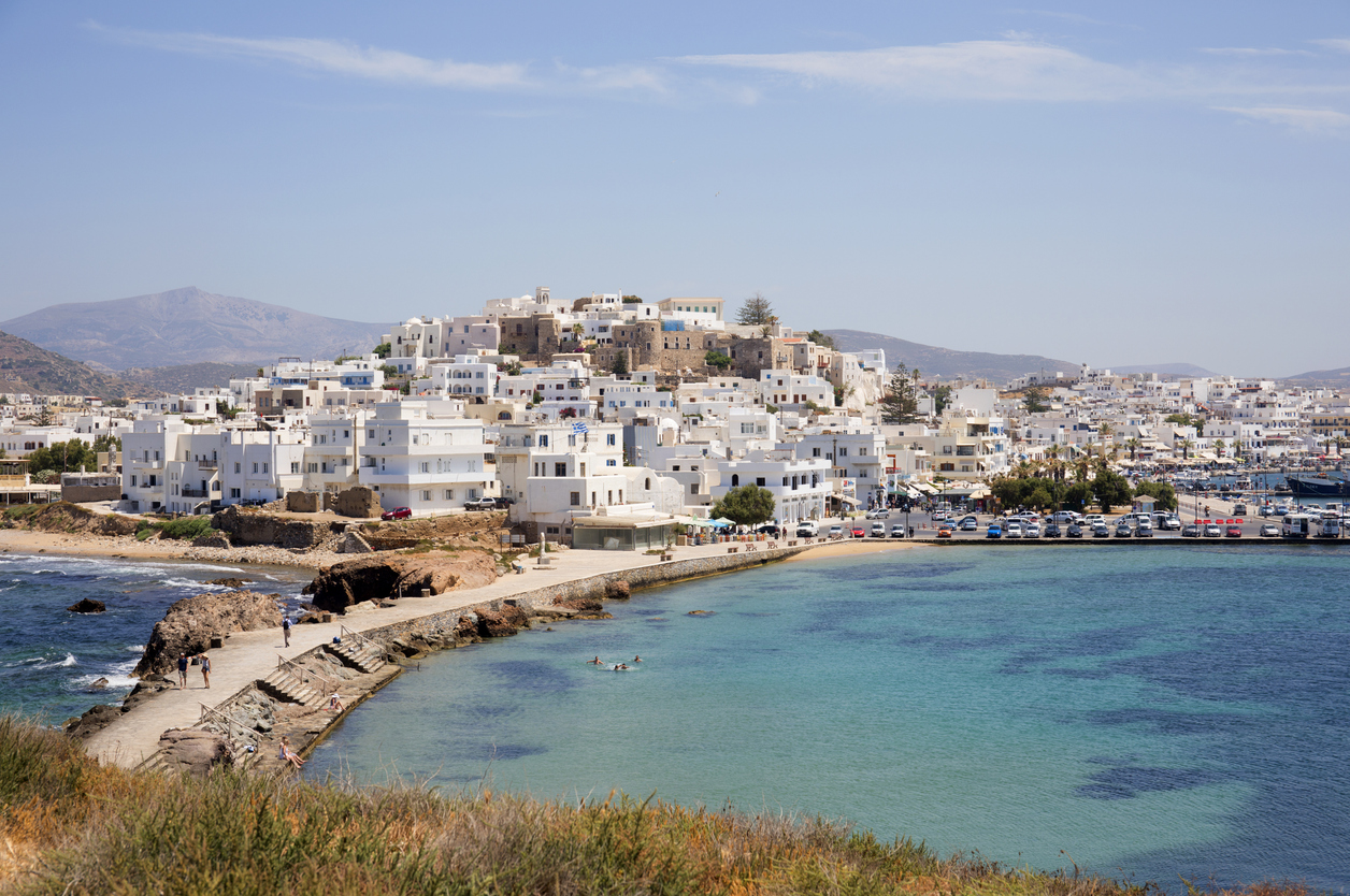 Panoramaaufnahme der Altstadt von Naxos in Griechenland mit weißen kubischen Häusern, engen Gassen, dem Hafen und türkisblauem Meer, im Hintergrund sanfte Hügel und Berge.