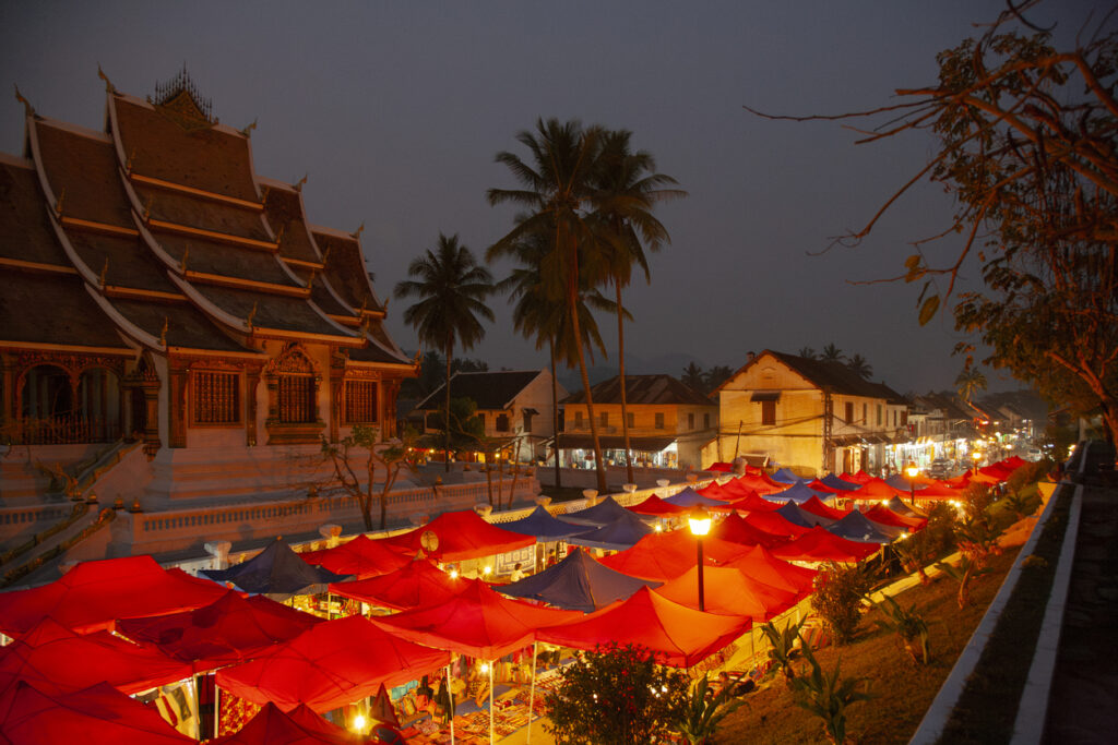 Abendaufnahme des Nachtmarktes in Luang Prabang, Laos, mit roten und blauen Marktzelten, beleuchtet von warmem Licht, im Vordergrund der prächtige Tempel Haw Pha Bang und Palmen im Hintergrund.
