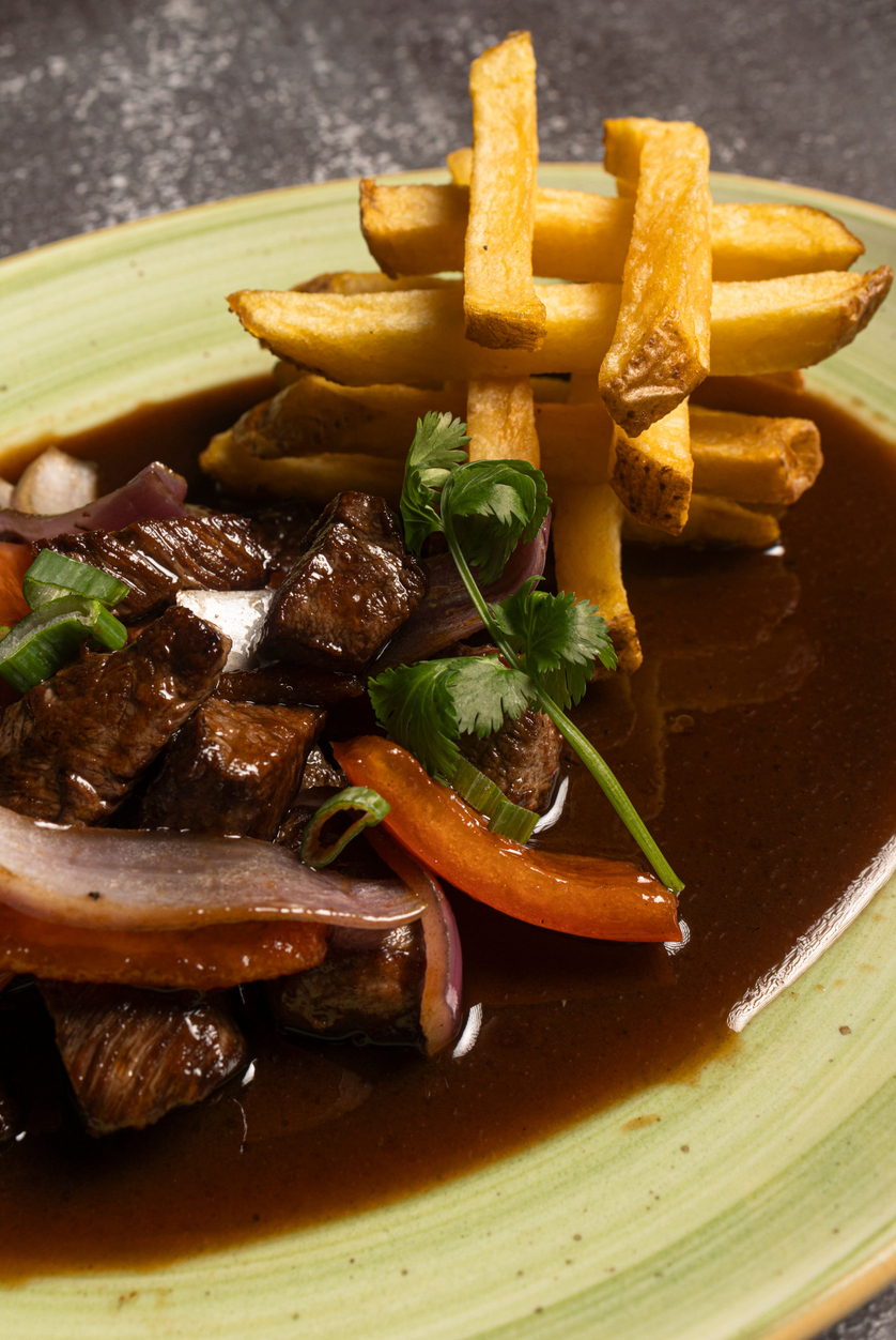 Nahaufnahme von Lomo Saltado, einem traditionellen peruanischen Gericht aus gebratenem Rindfleisch, Paprika, Zwiebeln und Koriander, serviert mit knusprigen Pommes frites auf einem grünen Teller.