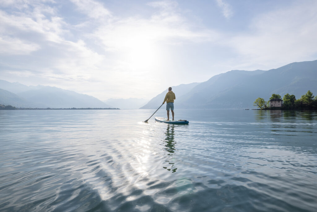 Person steht auf einem SUP-Board und paddelt auf einem ruhigen See, umgeben von Bergen in der Schweiz.