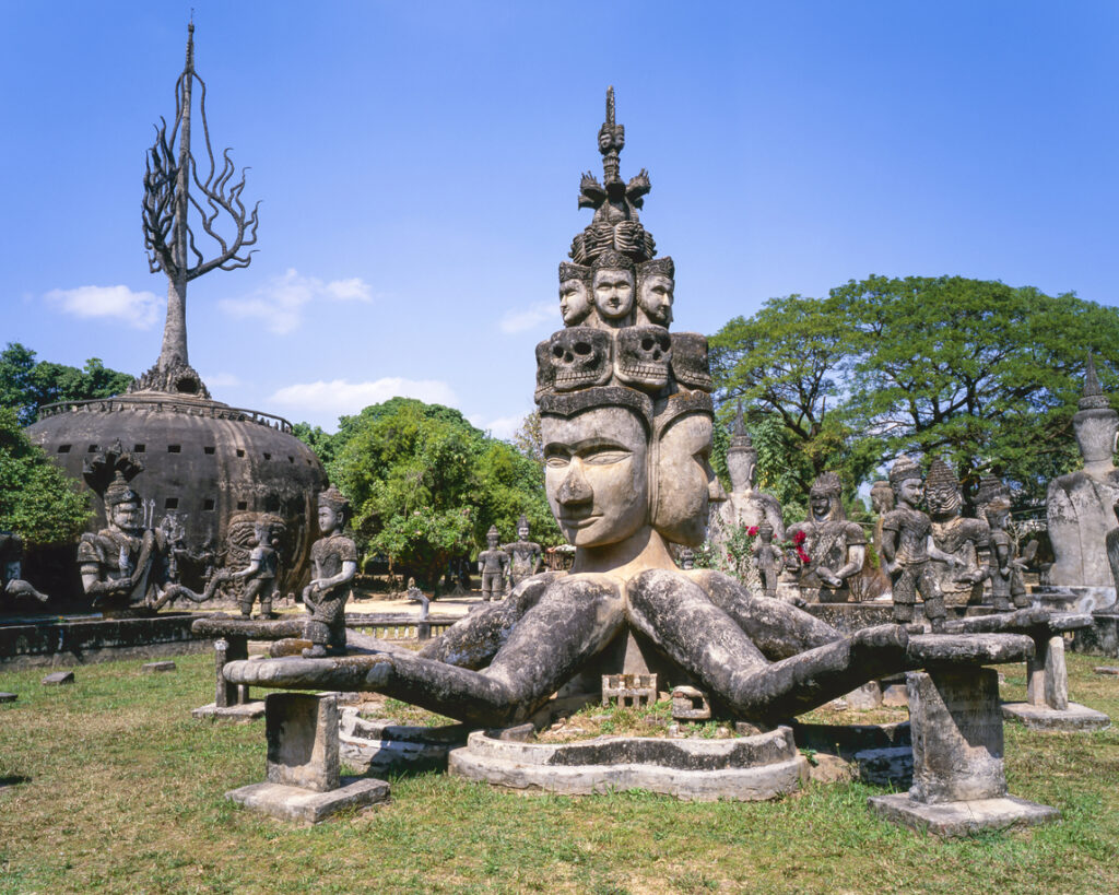 Große Steinskulptur mit mehreren Gesichtern und ausgestreckten Armen im Buddha-Park in Laos, umgeben von weiteren buddhistischen und hinduistischen Figuren unter blauem Himmel.