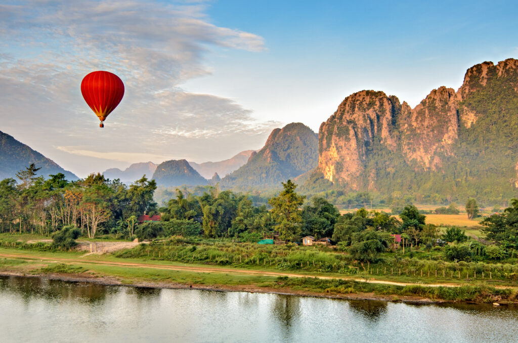 Roter Heißluftballon schwebt über der Flusslandschaft von Vang Vieng in Laos, umgeben von grünen Feldern, Karstbergen und Morgenhimmel.