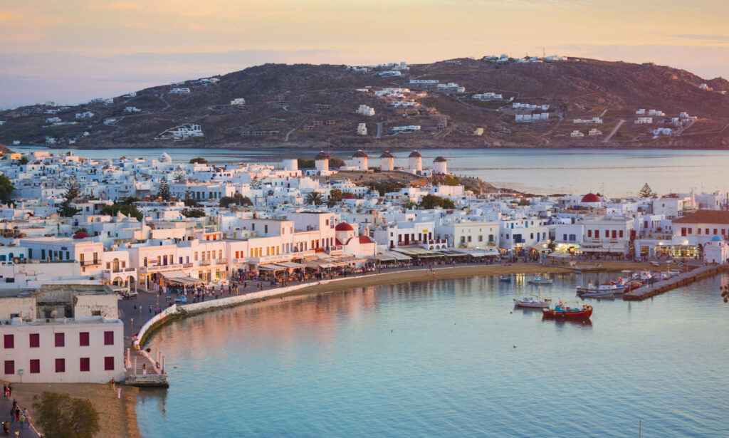 Panoramablick auf Mykonos-Stadt in Griechenland bei Sonnenuntergang, mit den ikonischen Windmühlen auf einem Hügel im Hintergrund, weißen Häusern mit blauen Akzenten, Uferpromenade, kleinen Fischerbooten im Hafen und sanft beleuchtetem Wasser.
