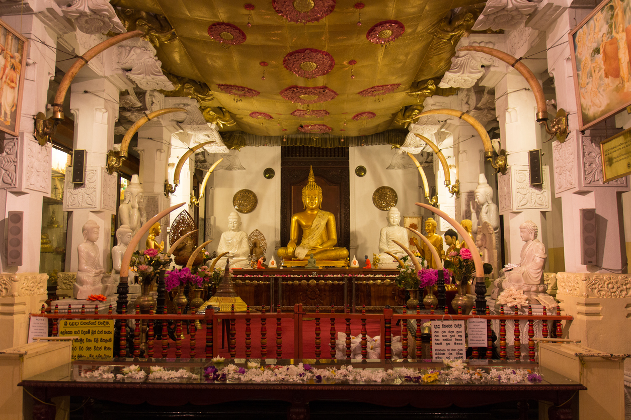 Innenansicht des Zahntempels in Kandy mit goldener Buddha-Statue, weißen Marmorfiguren und Blumenschmuck.