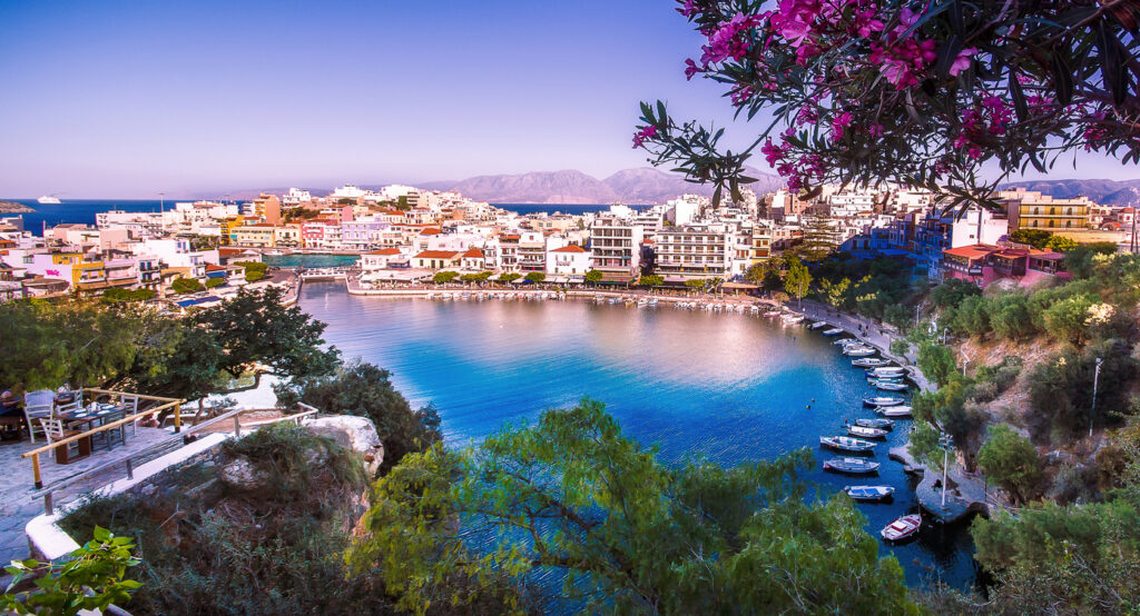 Panorama von Agios Nikolaos auf Kreta, Griechenland, mit dem malerischen Voulismeni-See, umgeben von bunten Gebäuden, Booten am Ufer, blühendem Oleander und Blick auf das Meer im Hintergrund.