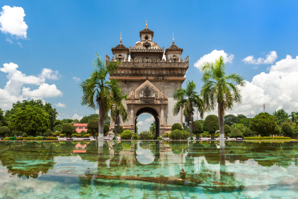 Das Patuxai Monument in Vientiane, Laos, mit kunstvollen Verzierungen, Palmen und seiner Spiegelung im Wasserbecken unter blauem Himmel.