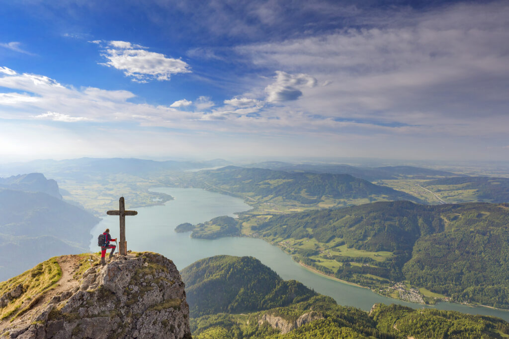 Bergsteiger steht am Gipfelkreuz des Schafbergs in den Alpen und blickt auf den Wolfgangsee, umgeben von grünen Hügeln und malerischen Tälern.