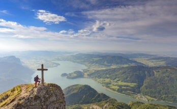 Bergsteiger steht am Gipfelkreuz des Schafbergs in den Alpen und blickt auf den Wolfgangsee, umgeben von grünen Hügeln und malerischen Tälern.
