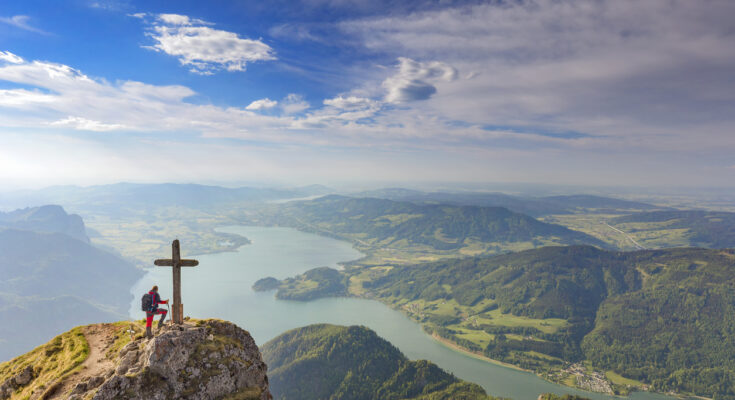 Bergsteiger steht am Gipfelkreuz des Schafbergs in den Alpen und blickt auf den Wolfgangsee, umgeben von grünen Hügeln und malerischen Tälern.
