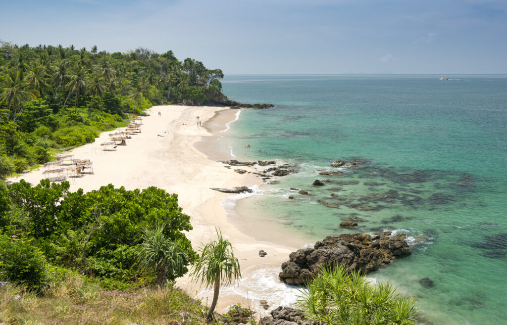 Tropischer Sandstrand auf Koh Lanta in Thailand mit grünem Dschungel, türkisfarbenem Wasser und Felsen am Ufer.