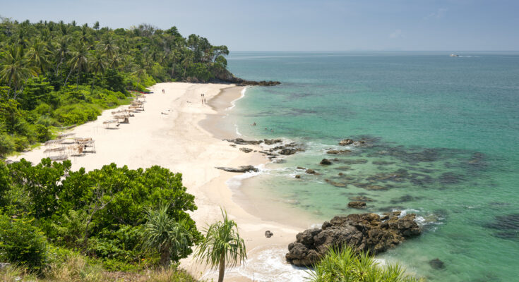 Tropischer Sandstrand auf Koh Lanta in Thailand mit grünem Dschungel, türkisfarbenem Wasser und Felsen am Ufer.