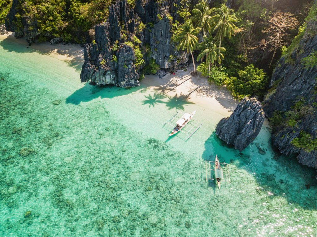 Luftaufnahme eines einsamen Strandes auf den Philippinen mit weißen Sand, Palmen, Felsformationen und traditionellen Auslegerbooten im klaren Wasser.